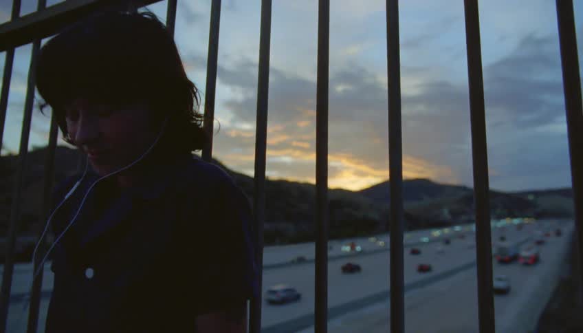 Girl with wavy hair and blue shirt leans against iron fences on highway overpass at dusk listening to music through earphones