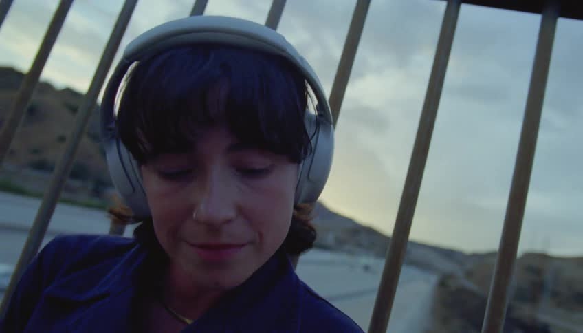 A teenage girl is sitting on a bridge leaning against the fence and listening to music in her headphones as traffic moves on the street below with mountains in the distance