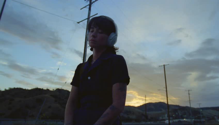 Woman with black polo shirt and tattoos listens to music through white headphones outdoors