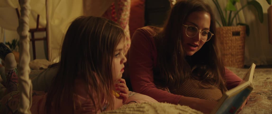 Mother with glasses brown wavy hair and red sweatshirt reading book to daughter lie down in living room