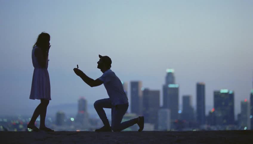 Man with cap t shirt and jeans proposing to woman with white dress and saying yes are silhouetted against the city skyline