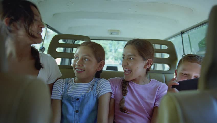 Woman talks to girls and boy playing smartphone game in the back of car