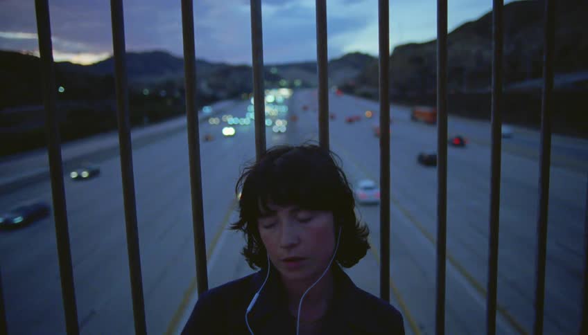 Woman with wavy hair and earphones leans against the iron fences and listens to music through earphones on highway overpass