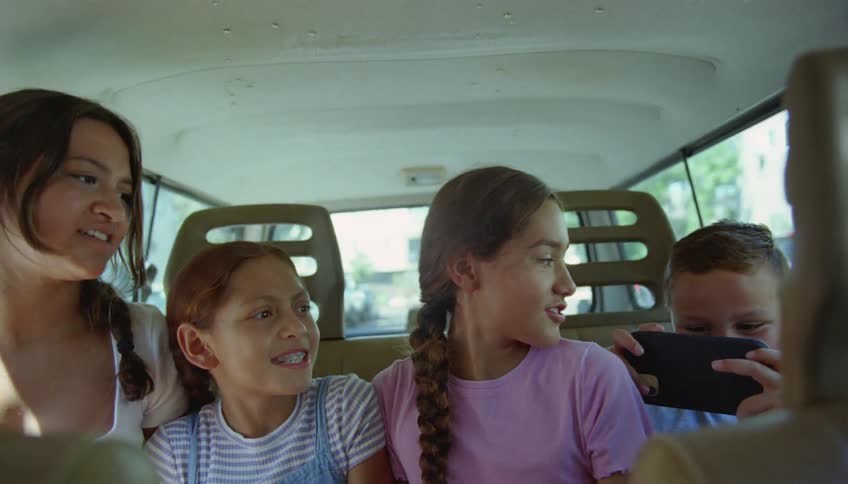 A boy sits in a vehicle next to his three sisters and plays on a cell phone as they fight and make faces at each other
