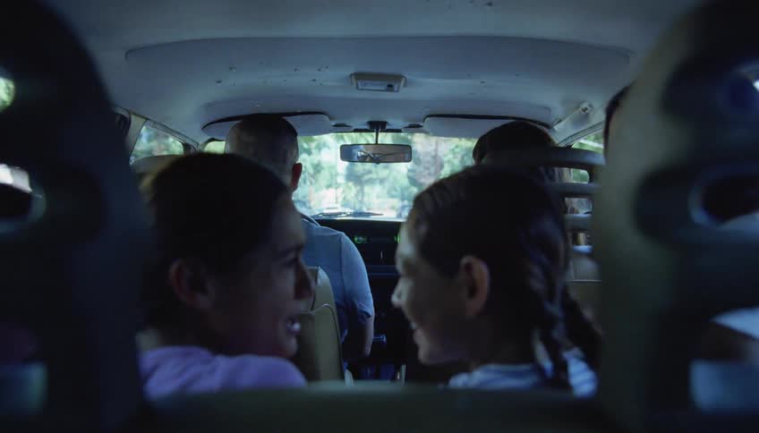 Young girls are talking and playing around as they ride in the back of a van as it travels on a road and their mother turns around and speaks to them