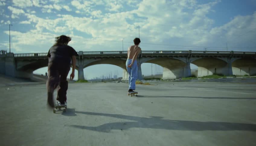 Two teenage boys skateboard on a road under a bridge that cars drive over
