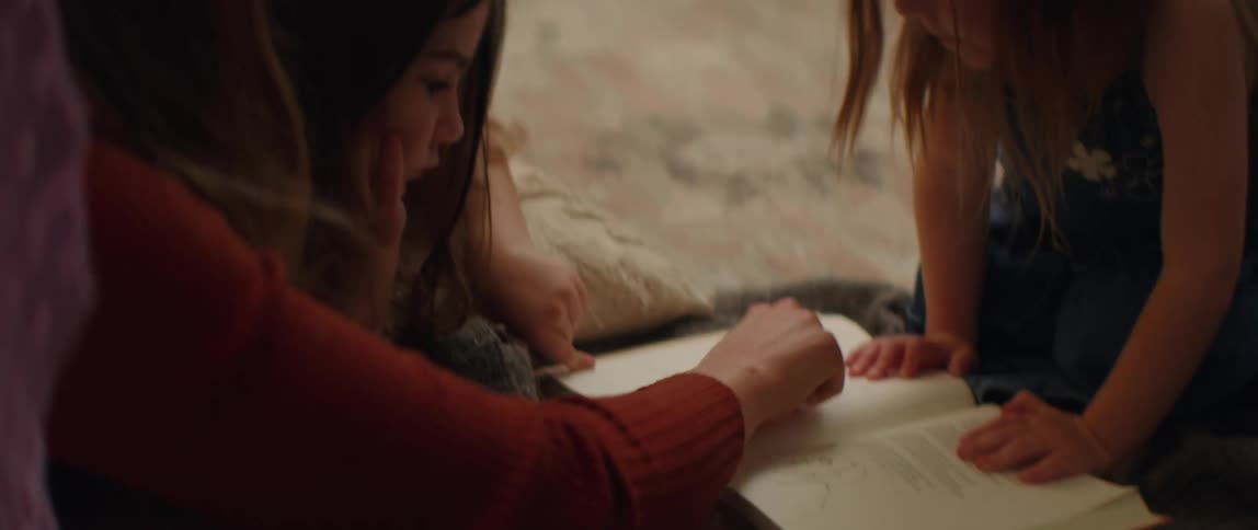 A mother lies on the floor with her two daughters and points at a book that they all are looking at