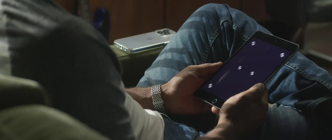 A man sits at home and holds a tablet while his cell phone rests on the couch next to him