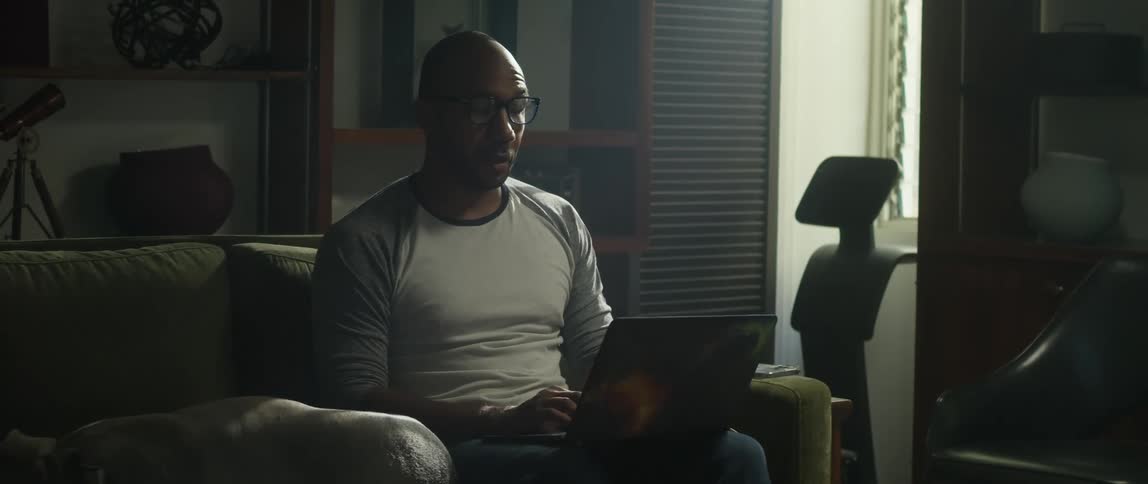 A man is sitting on the couch typing on a laptop computer and his dog is lying next to him
