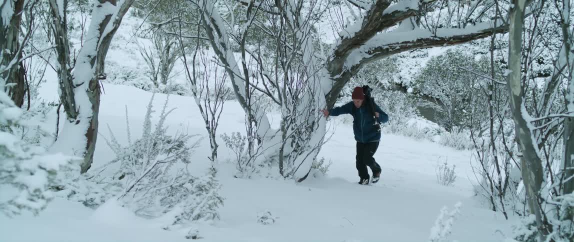 Man with red wooly hat and dark blue coat and pants carries tripod walks snowy uphill