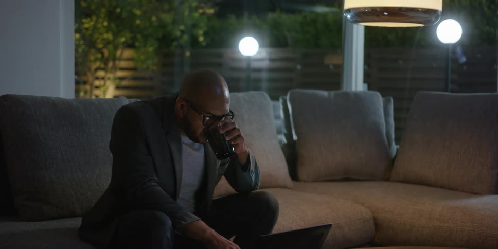 A man drinks from a cup while working on his laptop at home