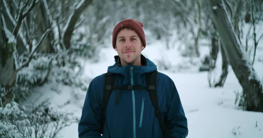 Young happy man stands relaxedly in front of a forest covered in snow looking at the camera