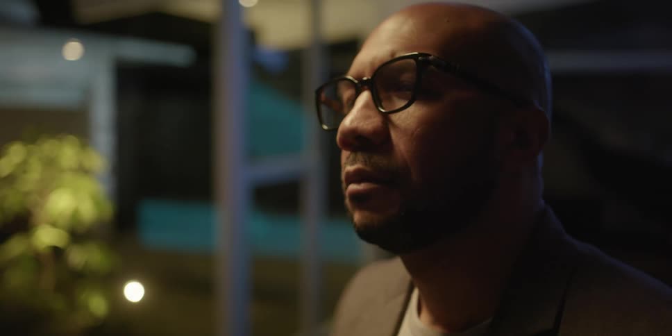 Bearded bald man wearing glasses steps into the living room of a luxurious house at night