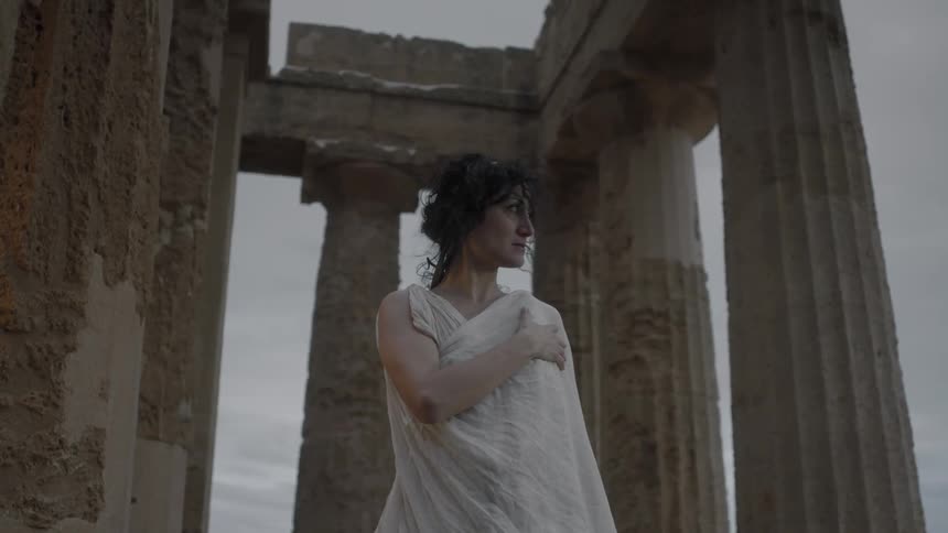 A woman in a diaphanous costume stands near some columns holding her arm across her chest