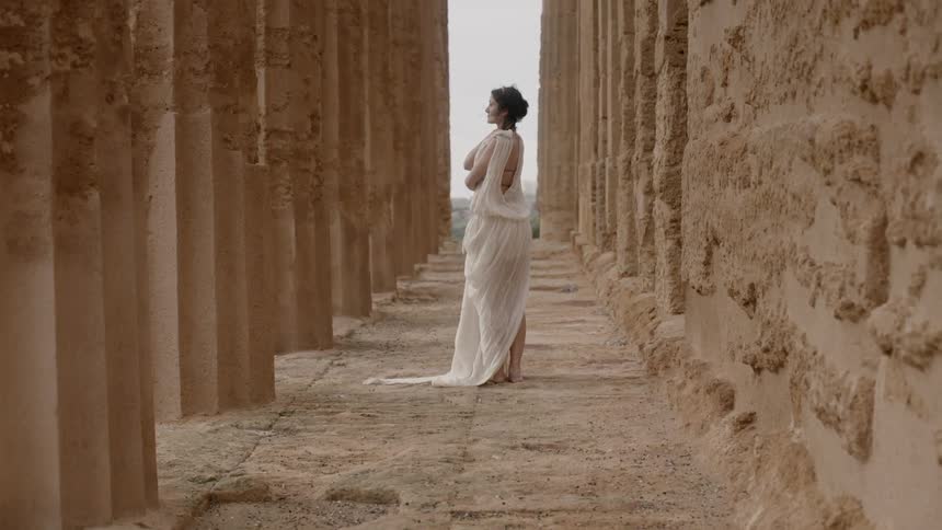 A woman wearing white stands behind large columns in the day time
