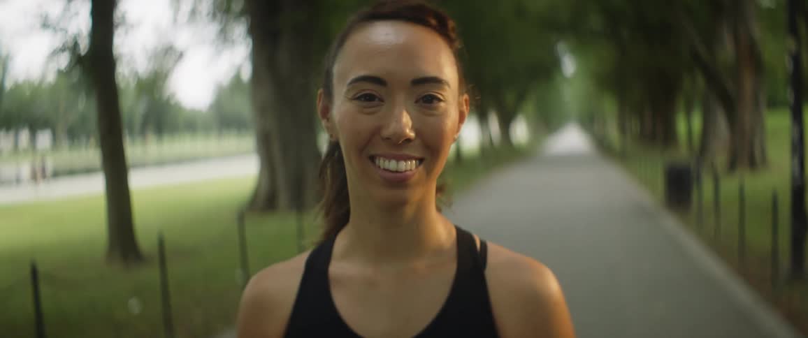 Woman wearing sports top stands smiling on path paralleling road as walkers walk along road in background