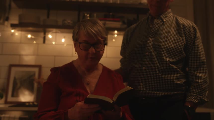 An elderly woman sits at a table with her son and picks up a book and starts to read and her husband comes and stands by her and puts his arm on her back
