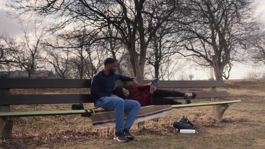 A woman lies down on her boyfriends lap to read a book on a bench in a park