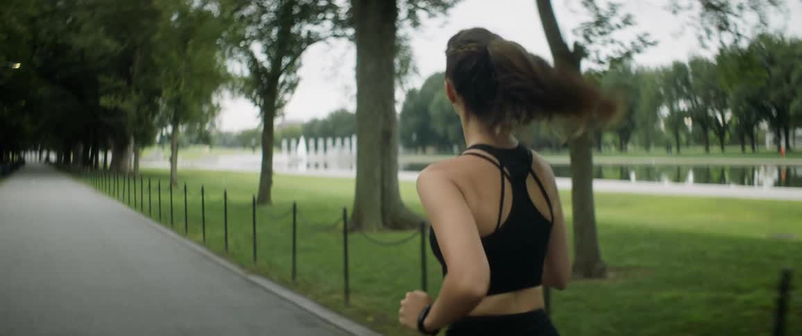Woman with ponytail sports bra and yoga pants jogs on road next to grass in public park