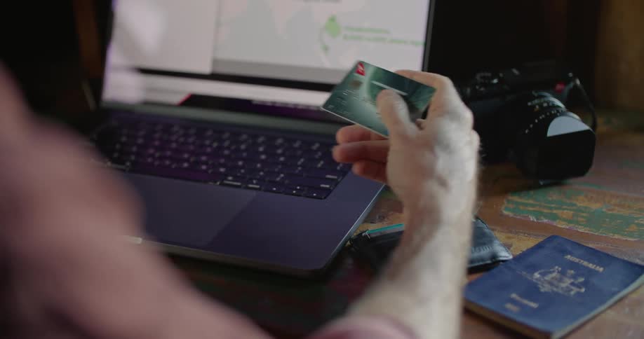 A man is holding his credit card as he types on a laptop computer and his wallet and passport are on the table