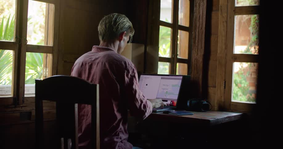 Man sitting at wooden table typing on laptop stops briefly to check their phone