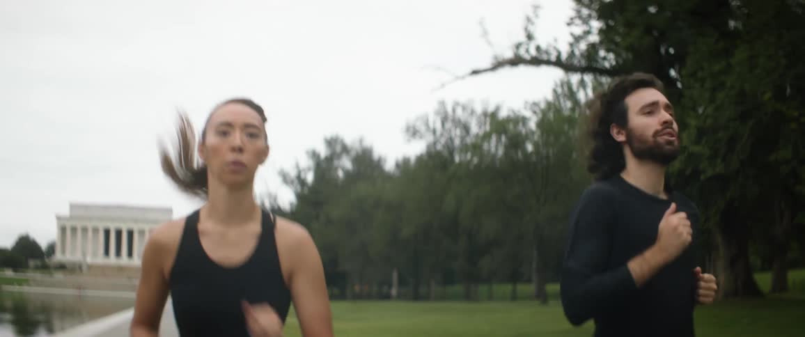 Woman with ponytail black sports bra and man with long wavy hair and black sweater jogs by lincoln memorial reflecting pool