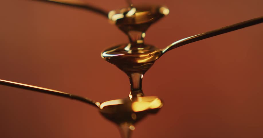 Thick amber honey dripping down on spoons over a brown backdrop