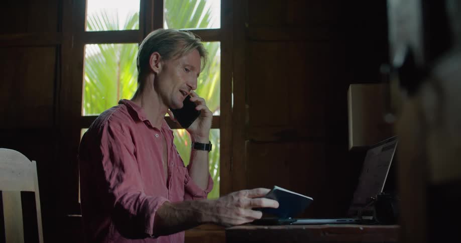 A man is sitting at a table with a laptop computer and talking on his phone as he holds a small book in his hand