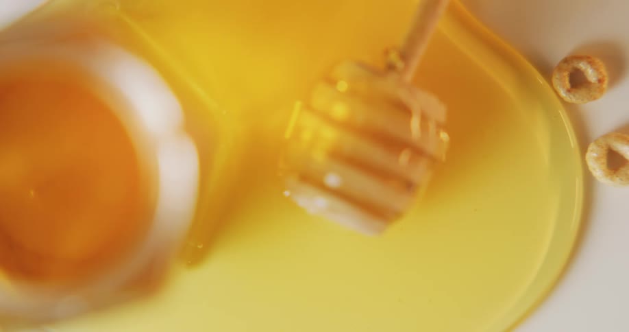 Puddle of honey spreads out from jar and honey dipper to touch cereal pieces