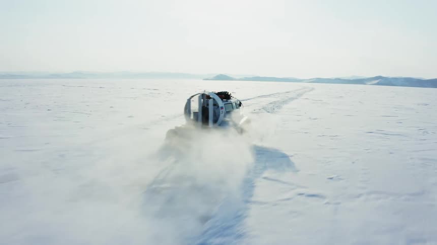 Aerial view of a hovercraft driving over the snowy lake on an arctic landscape near the mountains
