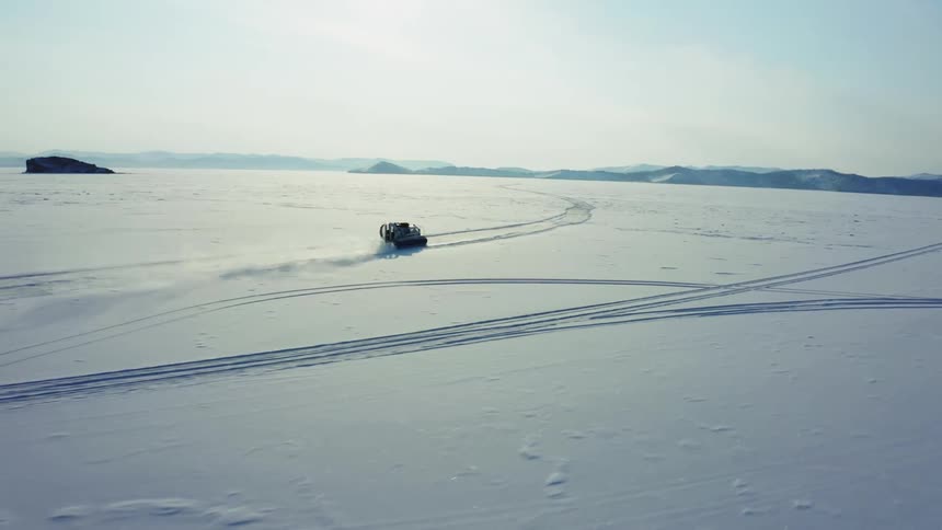 Large hovercraft moves across snow covered plain kicking up trail of snow with hazy mountains on the horizon