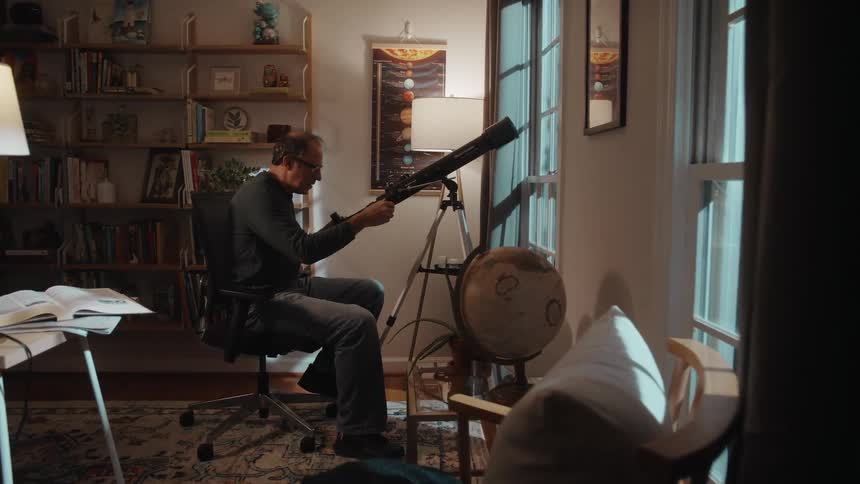 A man adjusts a telescope pointing out of a window of a house