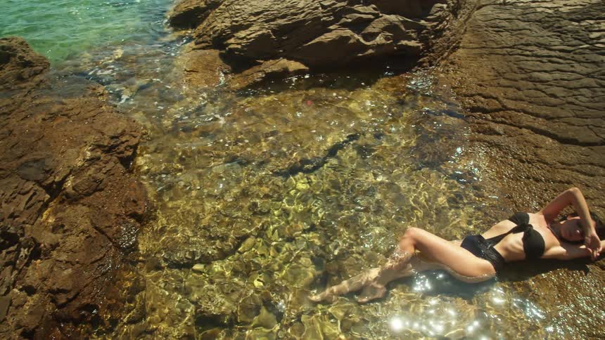 A woman in a swimsuit rests halfsubmerged atop a rock formation on the coast during the day