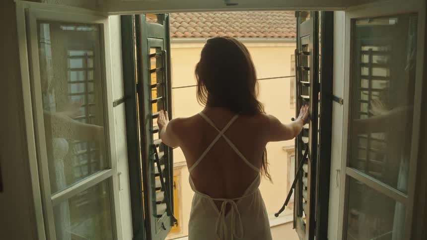 Young woman wearing a white dress opens the window shutters to look outside