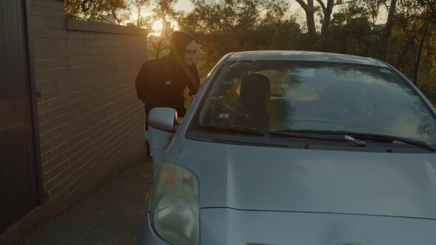 Male thief with black hoodie backpack and jeans breaks into car using poker at sunset