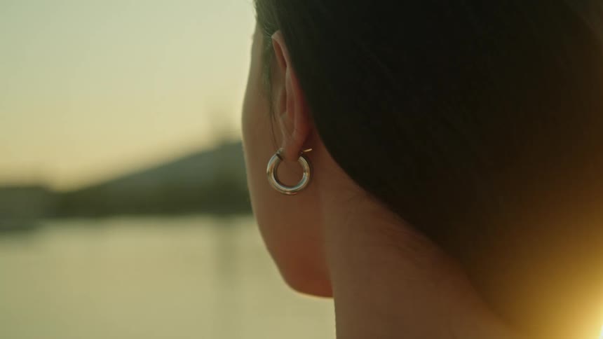 Woman with hoop earring and long dark hair watches sea at sunset