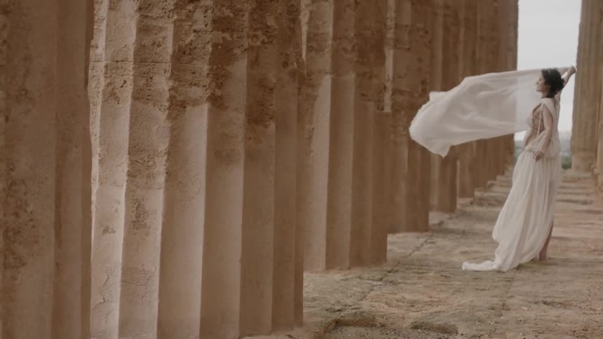 The wind blowing the train of a woman's dress around her as she stands next to columns at the acropolis