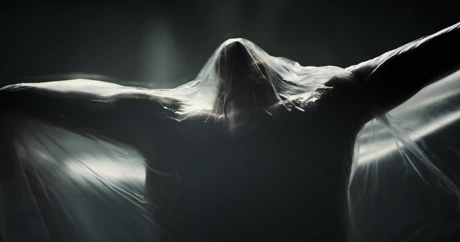 A woman rises her arms under a translucent plastic bag in front of a dark background