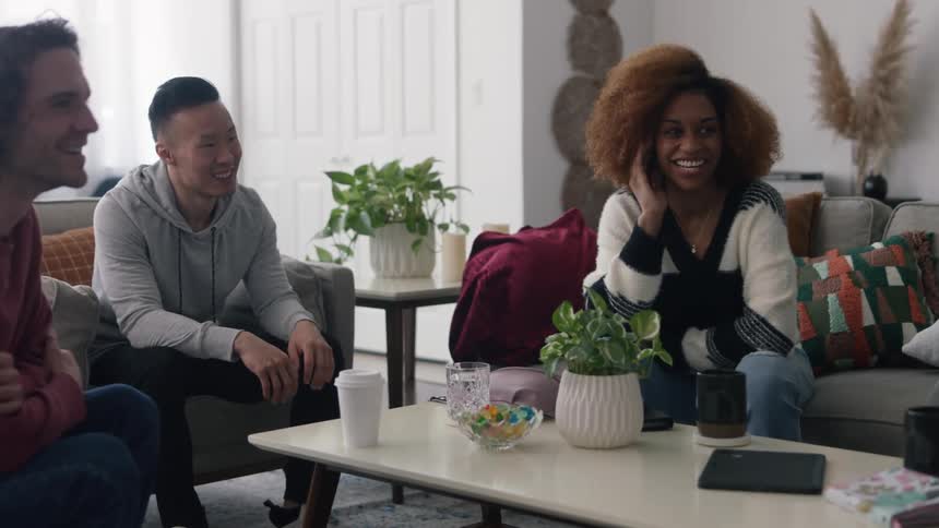 Woman with curly hair striped sweater and jeans sits on gray couch wit men wearing hoodie pants jeans and shirt in living room laughing