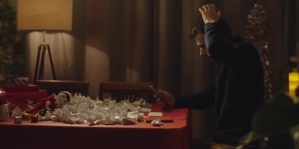 A man sits at home at a table with crumpled papers and picks up his cell phone and talks with someone as he runs his hand over his hair in frustration