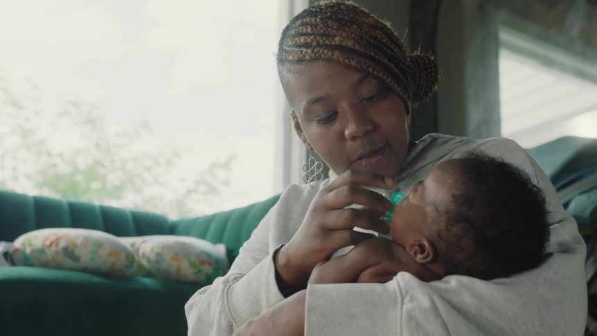 Mother with brown dreadlocks and gray hoodie puts pacifier in babys mouth in the living room at home
