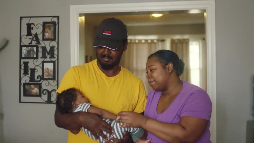 A father is holding his baby in his arms and the mother is admiring her child