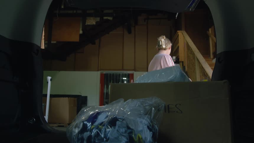 A woman loads boxes and bags into the back of a vehicle in a garage before closing the rear vehicle door