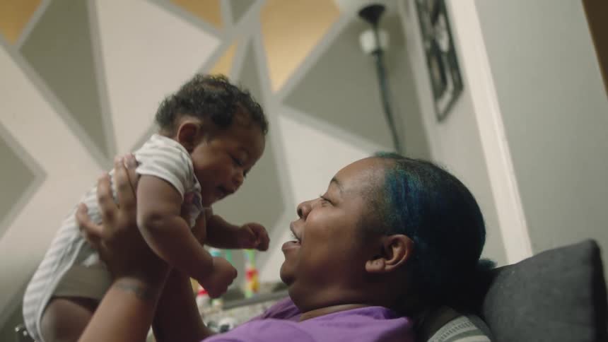 A woman is lying on the couch smiling and laughing as she lifts her baby into the air and bringing them down and rubbing noses