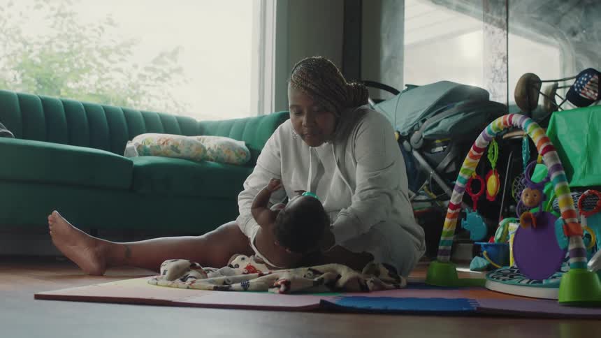 A woman sits on the floor at home holding her baby and gives him a kiss and lies him down on a blanket