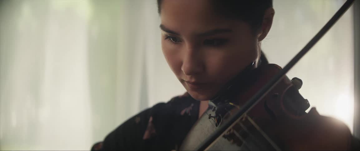 A woman looks serious as she plays the violin with sheers billowing in the breeze from open windows
