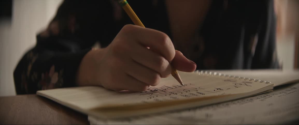 A womans hand writing sheet music notes in a notebook with pencil