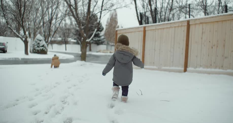 A young child walks through the snow in the front yard toward his sister