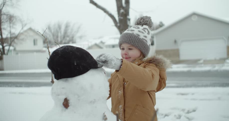 Girl wearing winter gear placescap onto snowman in snow covered neighborhood