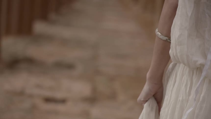 A woman wearing a diaphanous white dress walks among ruins
